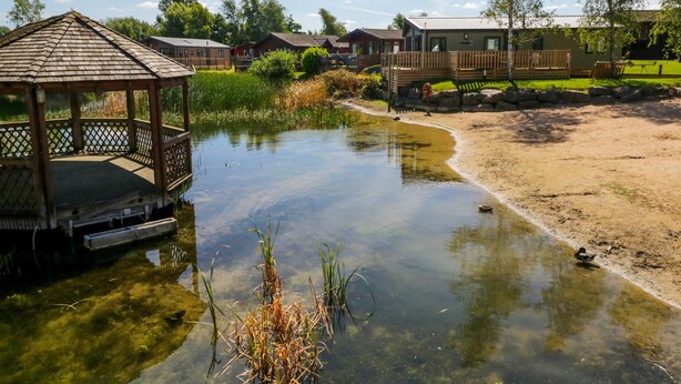 A tranquil lakeside setting with a wooden gazebo, a sandy beach, and a row of houses. - Lakeside 20 (Carnforth)