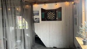 A room with a dartboard mounted on a wooden wall, a window with curtains, and a potted plant on a wooden table. - Sodra Gotland 14 bed With Pool (Havdhem)