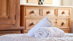 Un lit bien fait, avec une serviette blanche roulée, une commode en bois avec des tiroirs, et un vase avec des fleurs en arrière-plan.