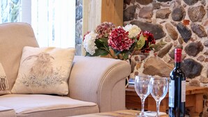 Un salon chaleureux avec un canapé beige, une table en bois sur laquelle sont posés des verres à vin et une bouteille, et un mur en pierre en arrière-plan.