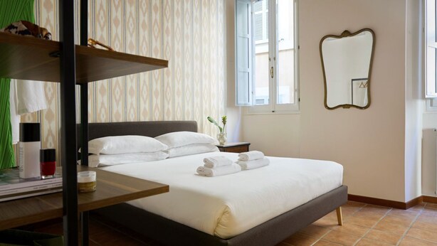 A neatly made bed with white linens, a bedside table with a glass, a mirror on the wall, and a window with blinds. - Trastevere Home - Santa Maria with beautiful view on the square (Roma)