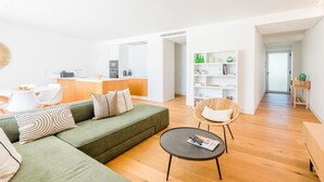 Un soggiorno moderno con un divano verde, un tavolino rotondo e una poltrona in vimini. Sullo sfondo si intravede una zona cucina con mobili bianchi e un piano di lavoro.