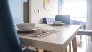 Ein Essbereich mit einem weißen Tisch, gedeckt für zwei Personen, einem blauen Stuhl und Blick ins Freie durch eine Glastür.
