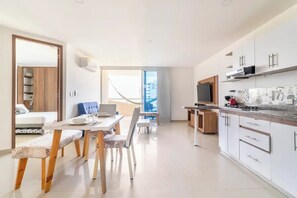 A modern kitchen with white cabinets, a built-in oven, and a microwave. There is a dining area with a table and chairs, and a large window providing a view of the city. - Oasis Apartment With Sea Views - Bello Horizonte (Santa Marta)