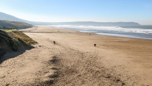 Una extensa playa de arena con olas suaves, cielo despejado y colinas a lo lejos.