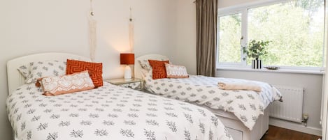 A bedroom with two beds, each with patterned bedding, a bedside table with a lamp, a hanging light fixture, and a window with a view of greenery.
