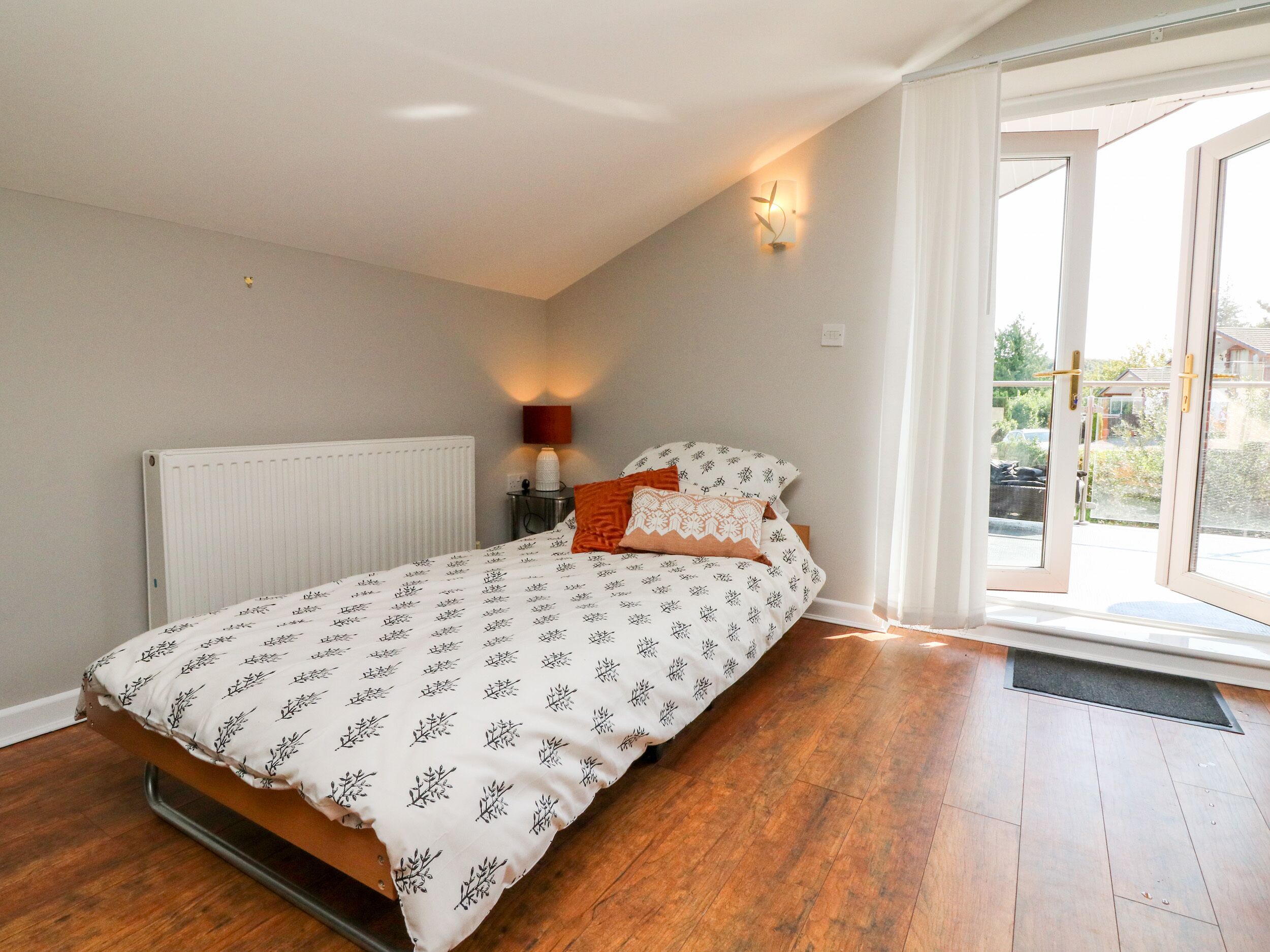 A bedroom with a bed, a radiator, a nightstand with a lamp, and a view of the outside through a door.