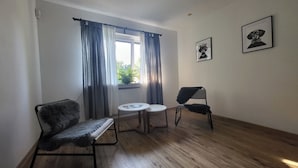 A room with a wooden floor, a chair, a small table, and a window with curtains. There are two framed pictures on the wall.