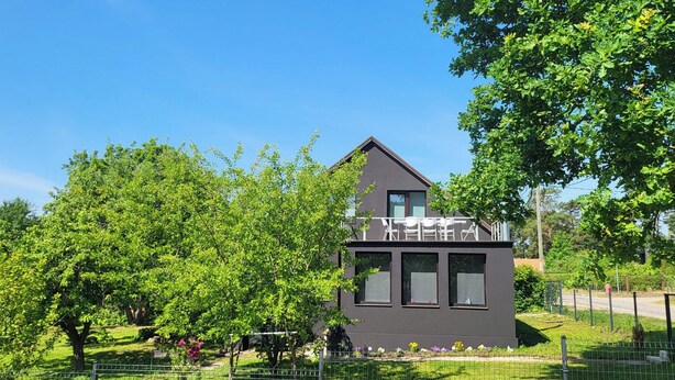 A modern two-story house with a balcony, surrounded by lush greenery and a fenced garden.