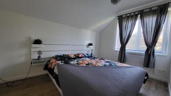Um quarto com uma cama, uma mesa de cabeceira com abajur, uma janela com cortinas e uma prateleira na parede com uma planta.