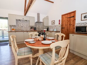 A dining area with a wooden table set for four, surrounded by chairs with plaid cushions. A kitchen with white cabinets and a tiled backsplash is visible in the background. - Shire Cottage (Driffield)