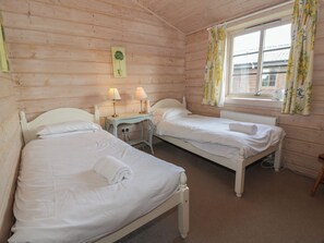 A wooden cabin room with two beds, a small table with lamps, a window with curtains, and a wall-mounted plant. - Lapwing Lodge (Dumfries)