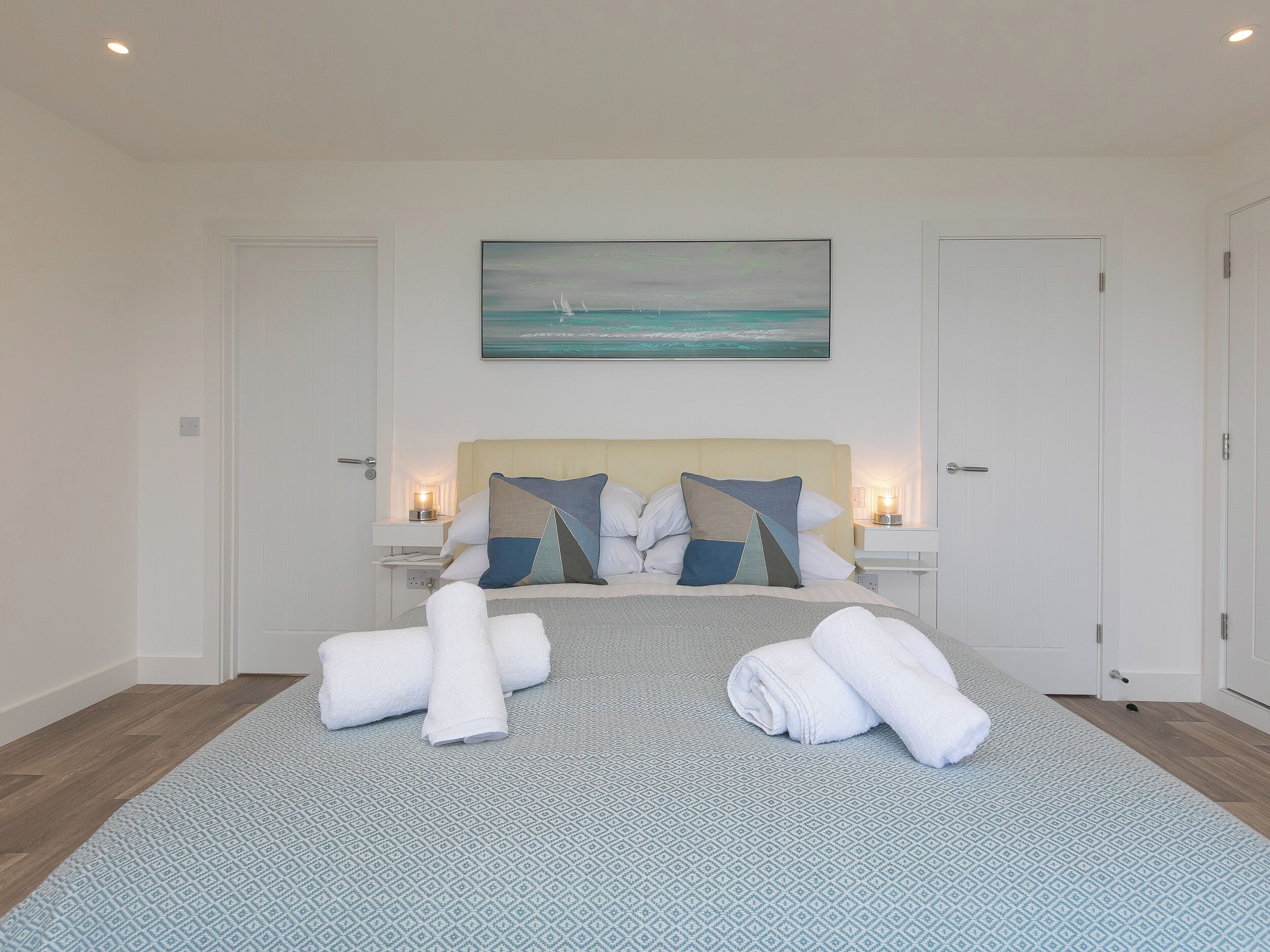 A neatly made bed with towels, a painting of the ocean, and a nightstand with a lamp.