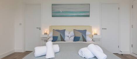 Un lit bien fait, agrémenté de serviettes, un tableau représentant l’océan et une table de chevet avec une lampe.