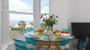 Uma sala de jantar com mesa e cadeiras de vidro, um vaso de flores e vista para o mar e casas.