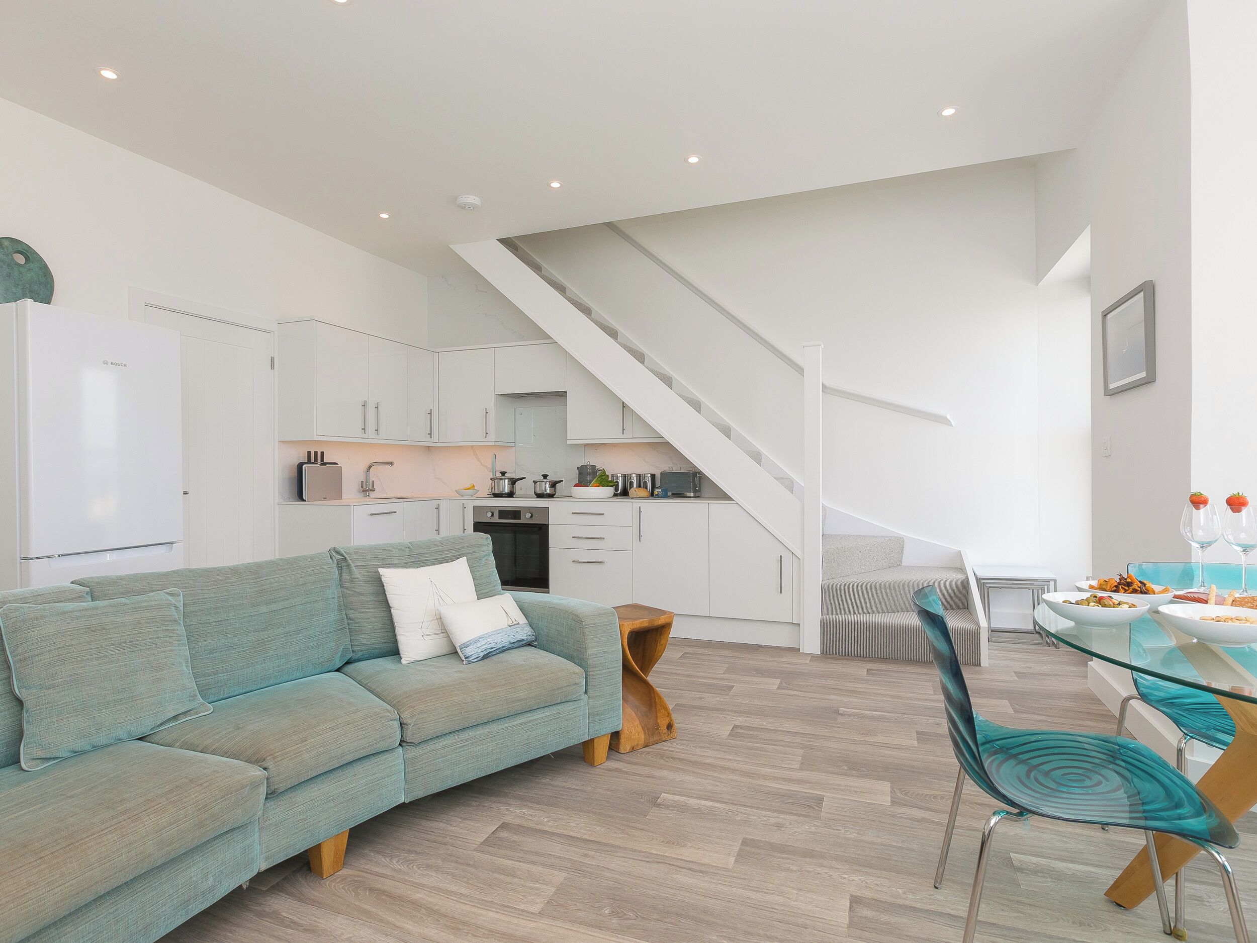 Une cuisine moderne avec des meubles blancs, un réfrigérateur Bosch et un coin repas comprenant une table en verre et des chaises de couleur turquoise.