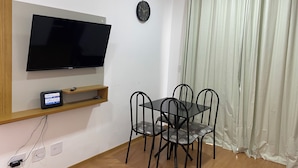 Une pièce dotée d’un téléviseur à écran plat fixé au mur, d’une horloge, d’une table à manger avec quatre chaises et d’une fenêtre avec des rideaux.