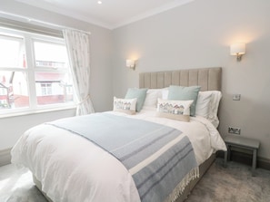 A bedroom with a large bed, a bedside table, a nightstand, a window with curtains, and a view of buildings outside.