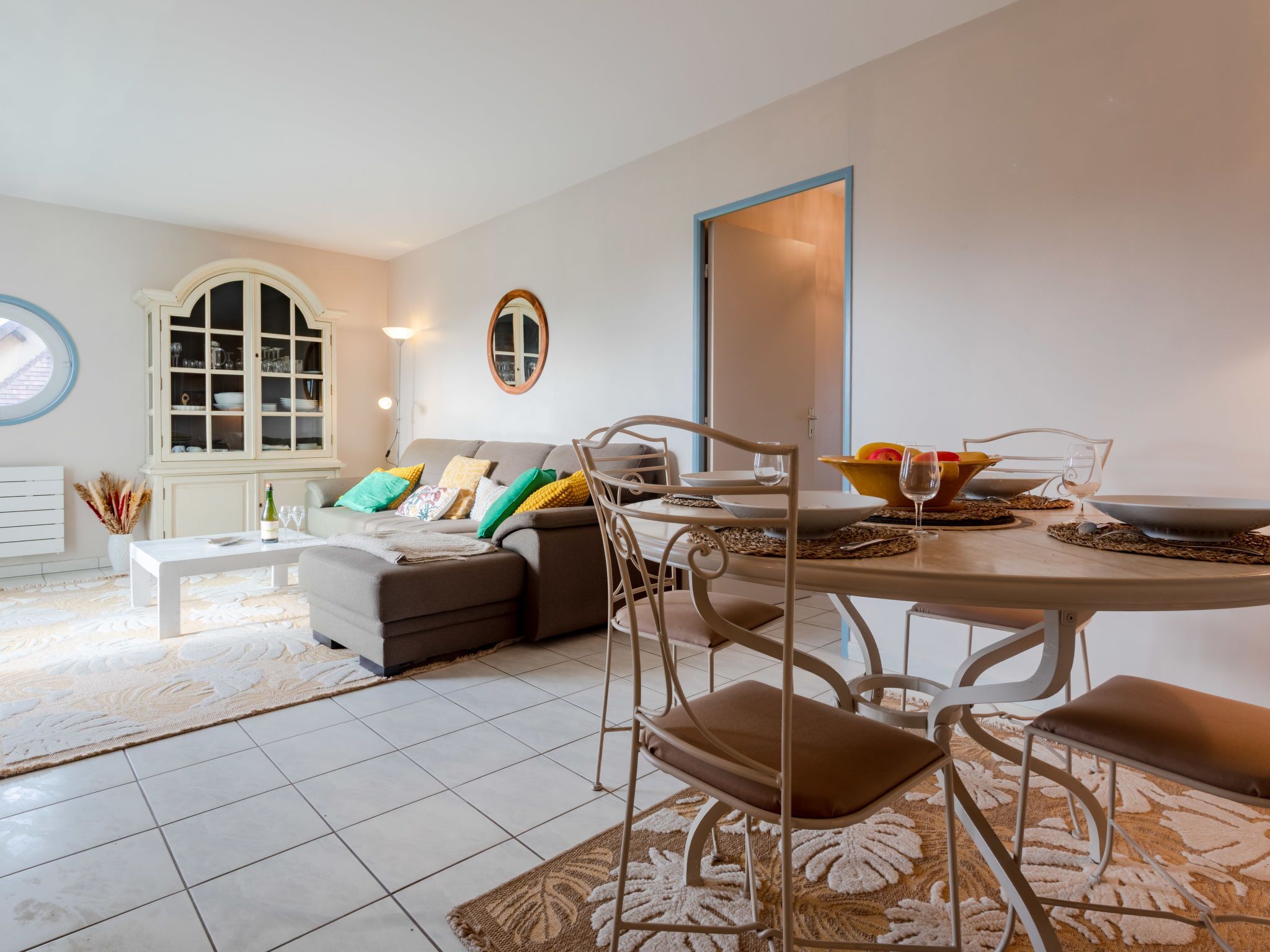 A modern living room with a sofa, dining table, and chairs. There is a window with a view, a cabinet, and a mirror on the wall.