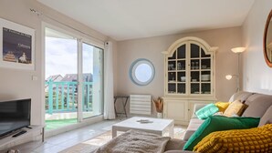 A living room with a sofa, a coffee table, and a TV. There is a window with a view of a balcony and a door leading outside. - Le Belvédère by Interhome (Cabourg)