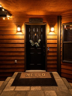 Eine Holztür mit einem 'WELCOME'-Schild darüber, ein Kranz hängt an der Tür und eine Fußmatte mit der Aufschrift 'HELLO' davor.