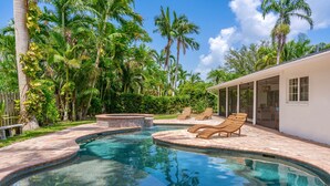 A swimming pool with a curved edge, surrounded by a brick patio. There are lounge chairs and a covered area with glass doors.