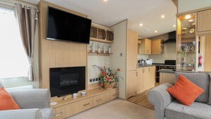 Un espacio habitable pequeño con televisión, chimenea, kitchenette y dos sofás grises con cojines naranjas.