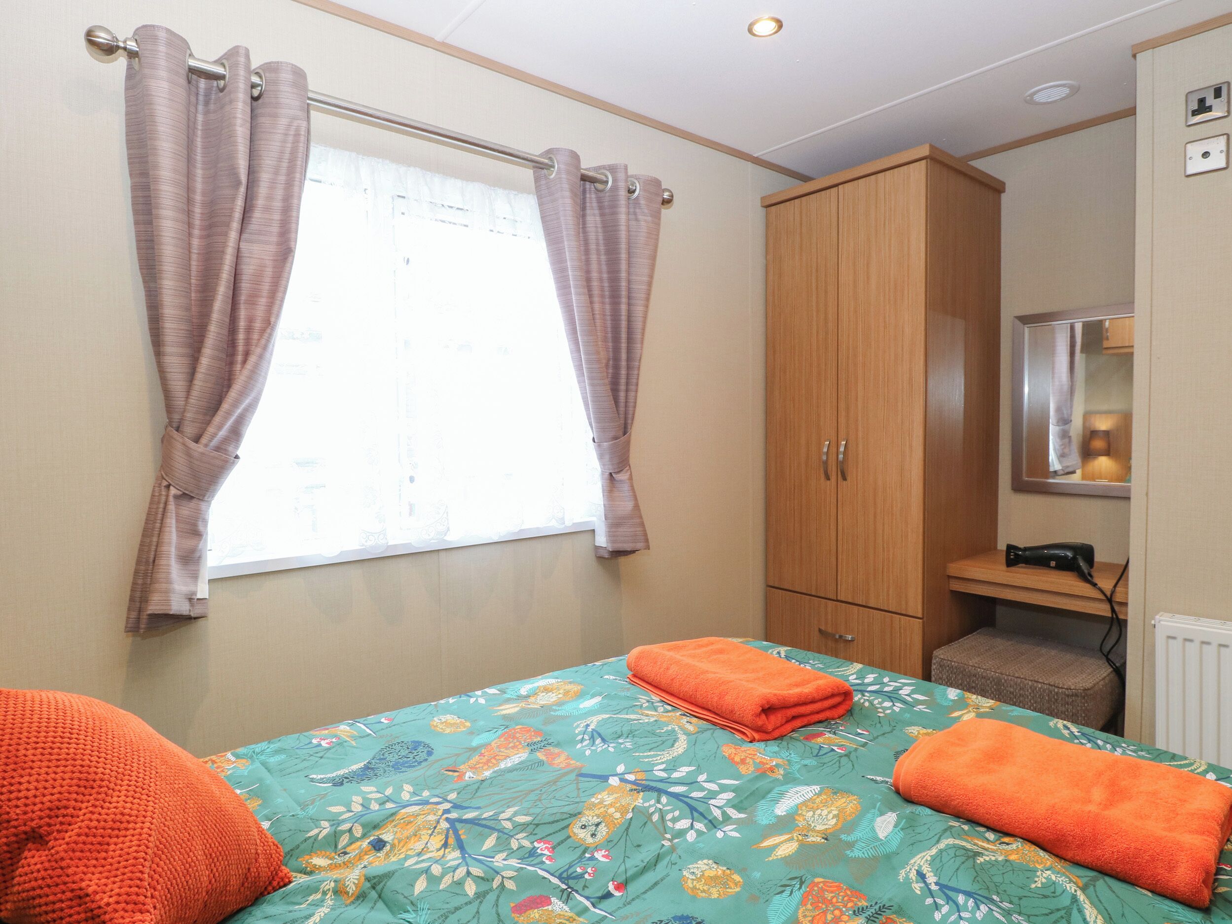 Une chambre à coucher avec un lit, deux serviettes orange, une armoire en bois, une petite table avec une lampe et une fenêtre avec des rideaux.