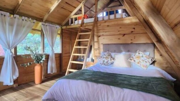 A cozy attic bedroom with a wooden ceiling, a bed with a green bedspread, a ladder, and a window with curtains.