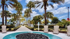 Un área de piscina con un jacuzzi circular rodeado de sillones de descanso, palmeras y un camino.