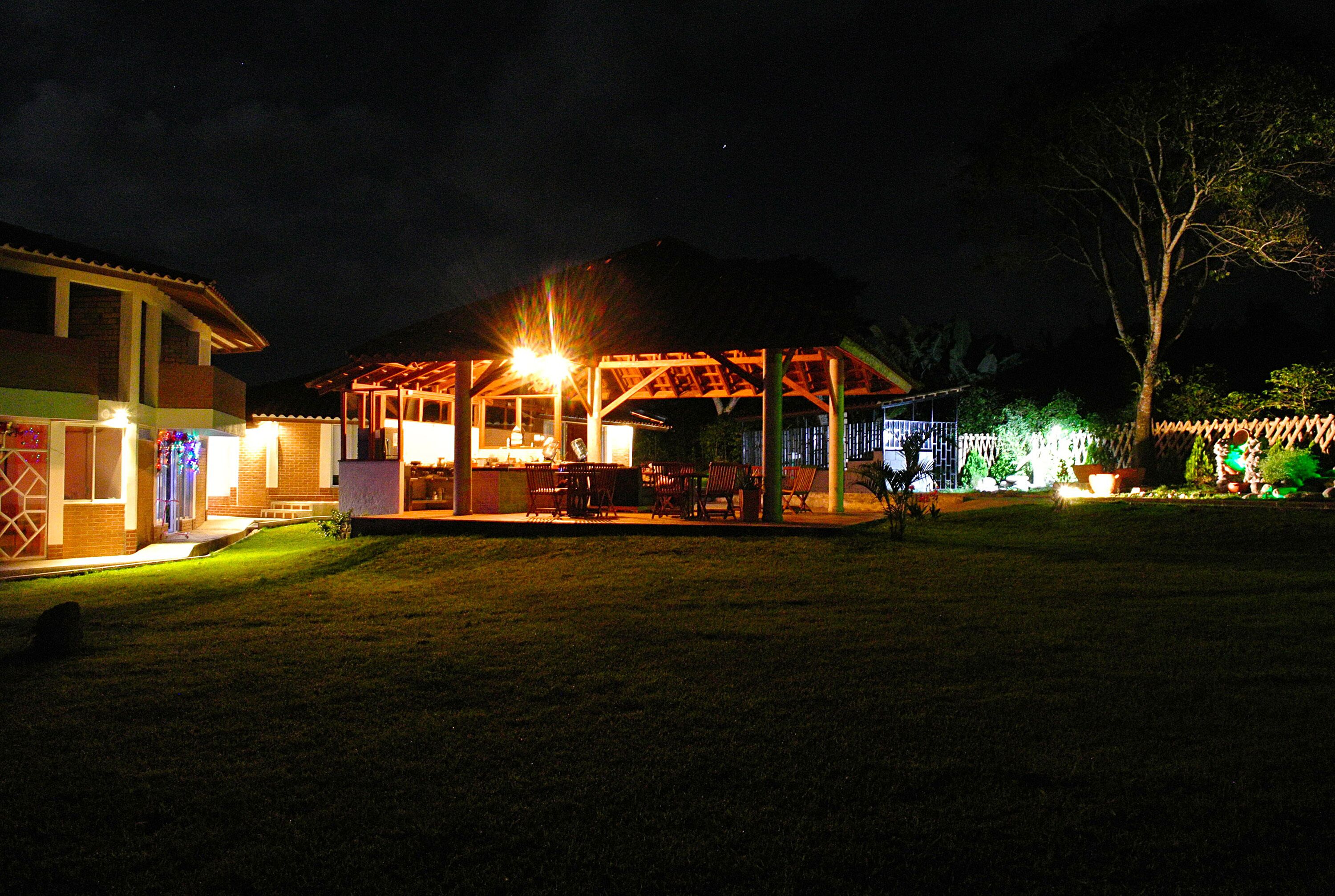 A covered outdoor area with tables and chairs, a building with a brick facade, and a tree in the background.