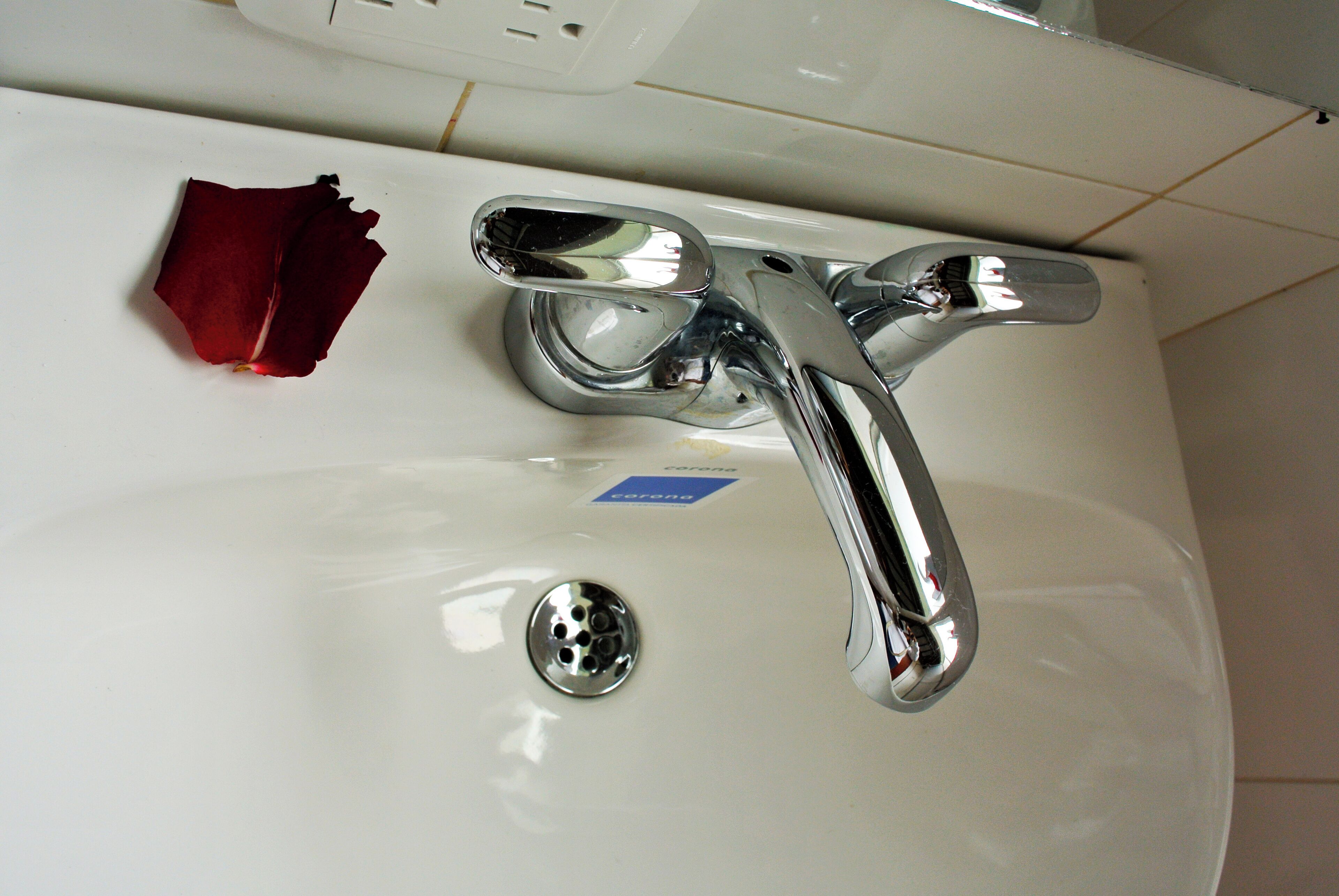Un lavabo blanc dans une salle de bain, avec un robinet chromé et un seul pétale de rose posé sur le comptoir.