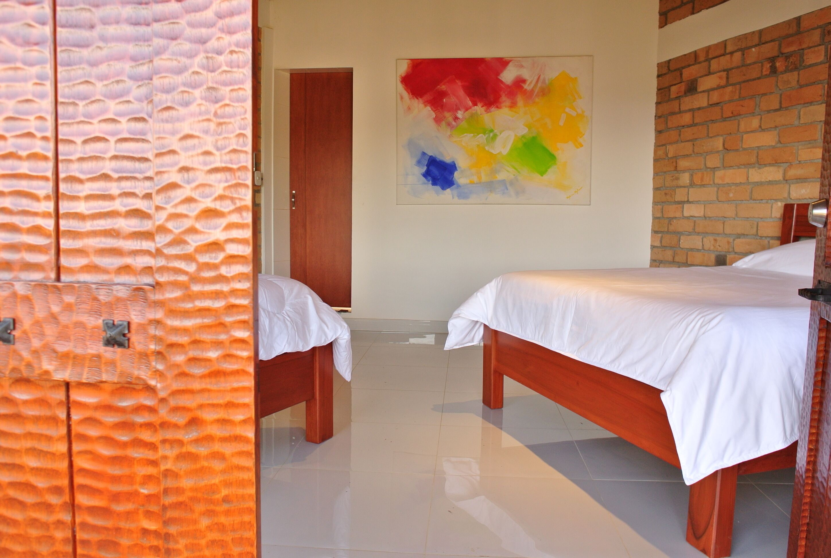 A hotel room with two wooden beds, a white wall with a colorful abstract painting, and a brick wall.