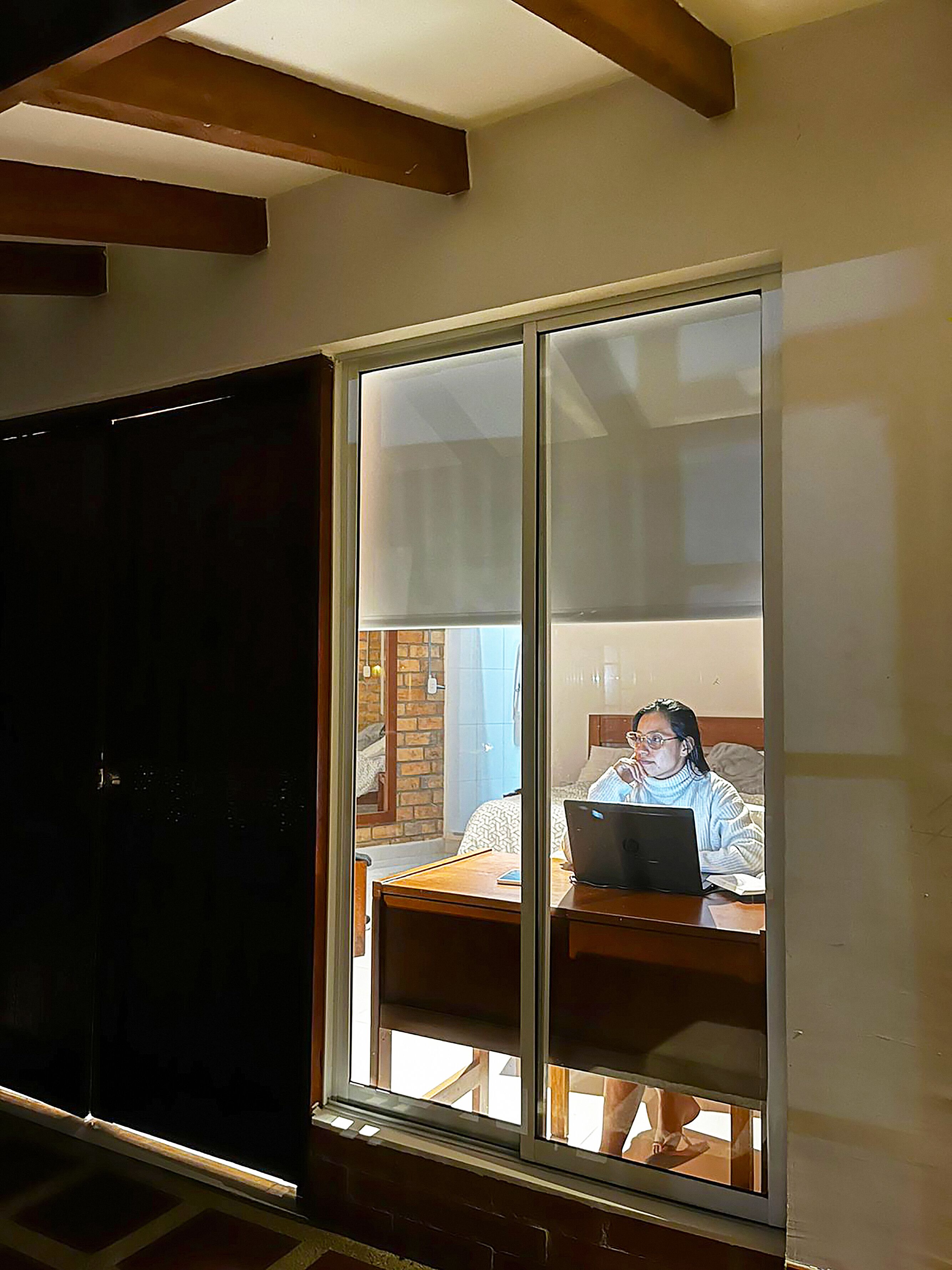 Ein Zimmer mit großem Fenster, einem Holztisch und einer Person, die einen Laptop benutzt.