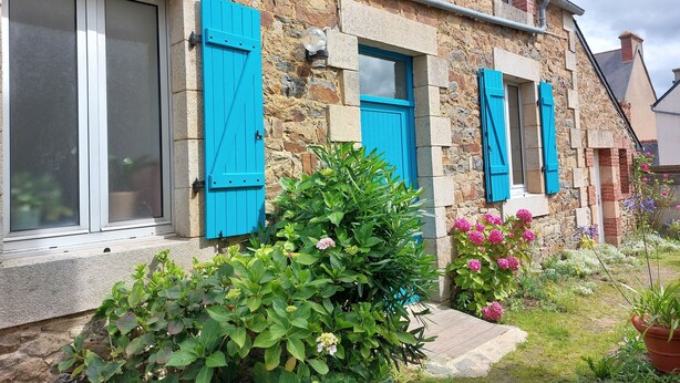 A stone building with blue shutters, a small garden with various plants and flowers, and a visible window. - Charmante Maison de Village , Tout à Pied, Commerces et Plage à 5 mn (Perros-Guirec)