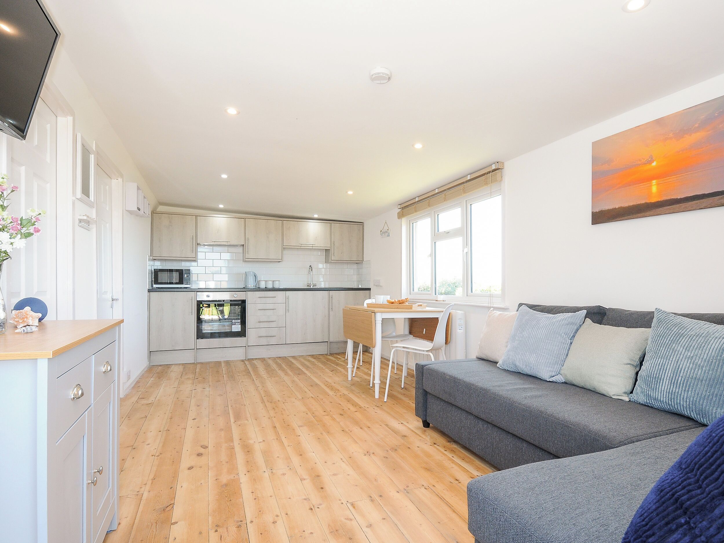 A modern kitchen with wooden cabinets, a dining area with a table and chairs, and a grey sofa in a living area with a large window.