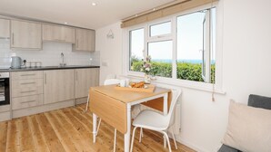 A modern kitchen with wooden cabinets, a dining table with chairs, and a view of the sea through a large window. - Lundy View Chalet (Bude)
