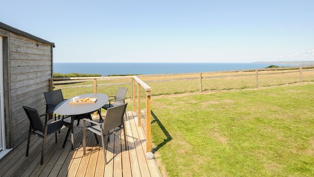 A wooden deck with a table and chairs overlooking a grassy field and the sea. - Lundy View Chalet (Bude)