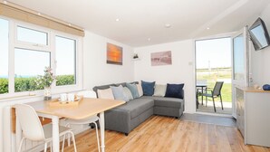 A modern living room with a grey sectional sofa, a wooden dining table with chairs, and a TV mounted on the wall. - Lundy View Chalet (Bude)