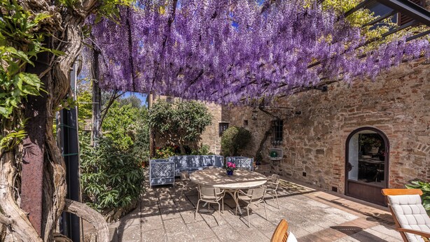 An outdoor patio with a stone wall, a round table with chairs, and a pergola covered in purple flowers. - Monticchiello (Pienza)