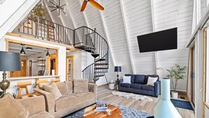 A spacious loft with a high ceiling, a wooden staircase, a large TV, and a cozy seating area with a coffee table and sofas. - NEW! A Frame Over Water - Sauna, Hot Tub, Views (Tahuya)