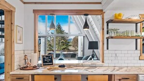 A modern kitchen with a large window, wooden bar stools, and a sign welcoming guests. - NEW! A Frame Over Water - Sauna, Hot Tub, Views (Tahuya)