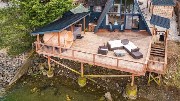 A modern house with a wooden deck, a glass conservatory, and a stone wall. - NEW! A Frame Over Water - Sauna, Hot Tub, Views (Tahuya)
