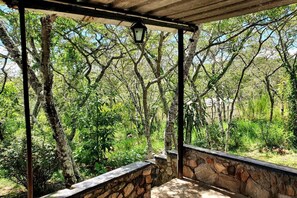 Un espace extérieur abrité, doté de murs en pierre et d’un plafond en bois, entouré d’une végétation luxuriante et d’arbres.