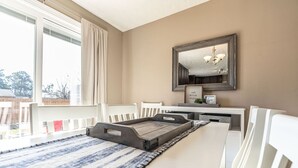 Un espace repas avec une longue table, des chaises blanches, un tapis à rayures bleues et blanches, un plateau en bois, un miroir et un lustre.
