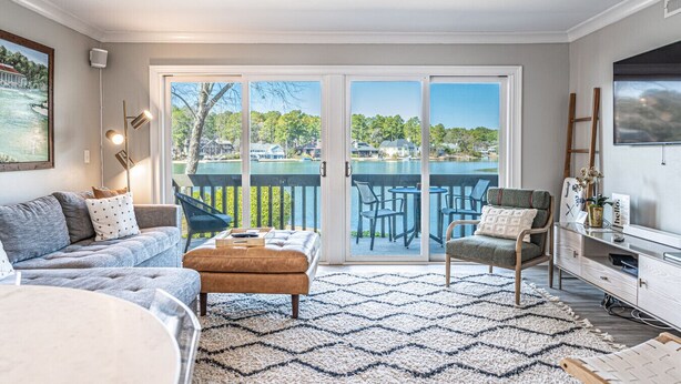 Un salon comprenant un canapé, un pouf et des fauteuils, un téléviseur, et une vue sur un lac et des maisons, visible à travers de grandes portes-fenêtres coulissantes.