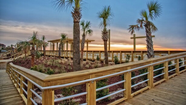 A wooden boardwalk with railings, palm trees, and a wooden structure in the background. - Canal-Front Townhome w/ Private Boat Slip, Pool & Beach Access (Carolina Beach)