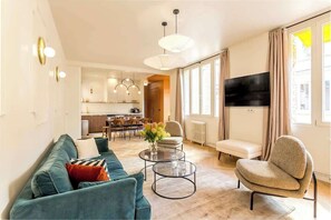 Una sala de estar moderna con un sofá, un sillón y una mesa de centro. Se observa una cocina al fondo con un comedor.