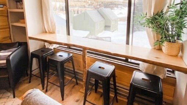 Interno accogliente di una baita con una grande finestra che si affaccia su case e montagne innevate. Si notano scaffali di legno, una scrivania e tre sgabelli.
