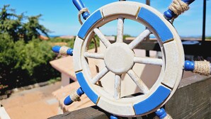 A close-up of a ship's wheel with blue accents and rope wraps, set against a clear sky and greenery. - Case Vacanza Rena Majore (Aglientu)